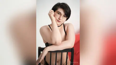 Young woman with short hair and glasses wearing black lingerie, posing thoughtfully in a bright indoor setting.