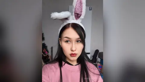 Young woman with bunny ears and pink shirt posing in a cozy indoor setting with a playful mood.