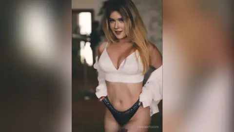 Blonde woman in a white crop top and Tommy Hilfiger underwear posing in a soft-lit indoor setting.