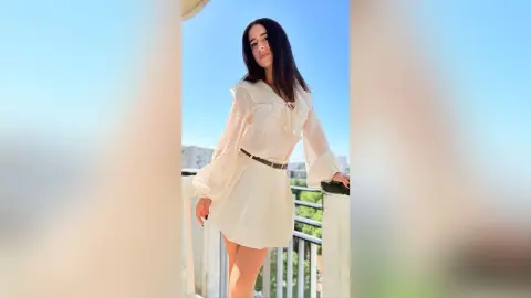 BellaLeen poses on a balcony wearing a white long-sleeved blouse and a short white skirt with a thin brown belt.