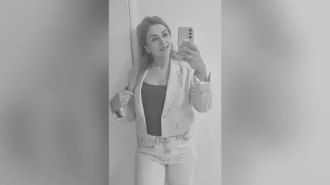 Jess poses in a light-colored blazer and matching jeans, holding a phone up to a mirror while looking at the camera.
