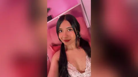 Young woman with long dark hair in a white lace top posing in a bright pink bedroom.