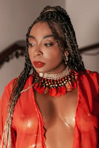 Beautiful Black woman with long braids wearing a red outfit and bold statement necklace in warm lighting.