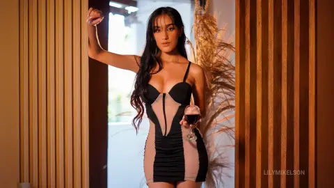 Elegant woman in a black cocktail dress holding a glass of red wine in a warm, moody indoor setting.