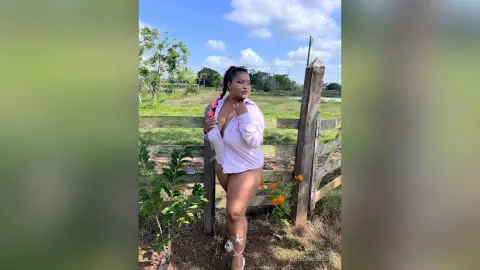 Curvy woman in a white button-down shirt posing confidently by a rustic wooden fence in a sunny field.