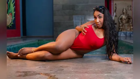 NaiomiLanne — Curvy woman in a red one-piece swimsuit relaxing by a stone fountain in a serene outdoor garden setting.