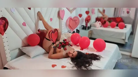 Romantic woman in red lingerie lying on a bed surrounded by red heart balloons and roses.
