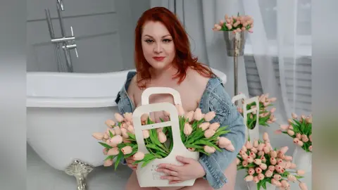 Redheaded woman in a denim jacket holding a bouquet of pink tulips in a bright, airy bathroom setting.
