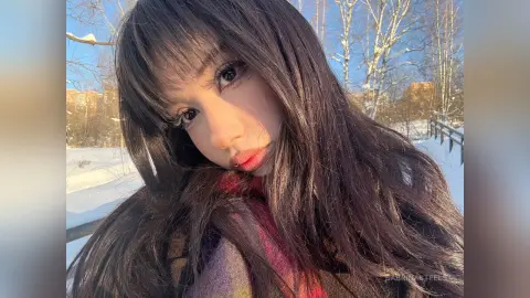 SabrinaSteels poses with long, dark hair and bangs against a bright, snowy outdoor background with trees and blue sky.