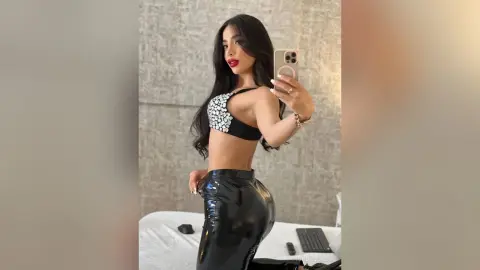 Young woman in black sequin crop top and shiny patent leather pants taking a mirror selfie in a modern bedroom.