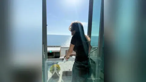 Woman in a black top and denim jeans standing on a balcony overlooking the bright blue ocean view.
