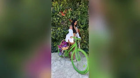 Smiling woman in a white shirt and shorts with a flower basket on a green bicycle against a lush garden wall.