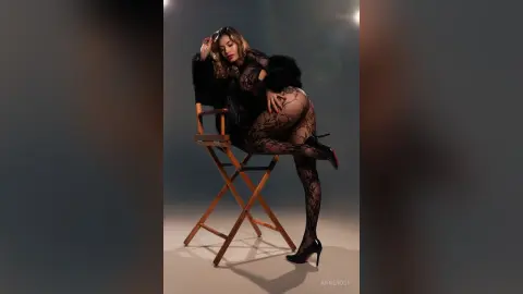 AnneRoss sits on a wooden chair wearing black lace lingerie and sheer black stockings with black high heels.
