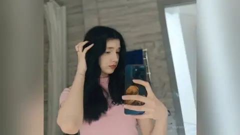 Young woman with long black hair in a pink t-shirt taking a mirror selfie in a modern bathroom.