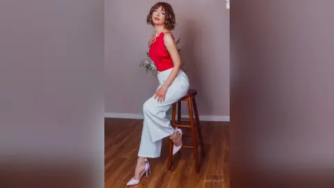 Stylish woman in a red sleeveless top and white wide-leg trousers sitting on a stool in a minimalist studio.
