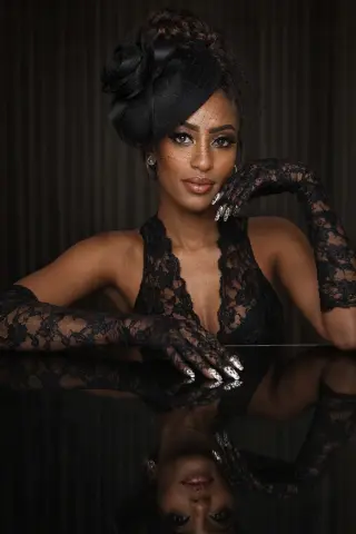 Elegant woman in black lace dress and floral fascinator posing glamorously against a dark background.