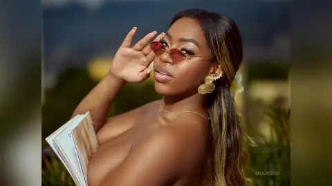 Glamorous woman in sunglasses and gold earrings reading a book in a sunny outdoor garden setting.