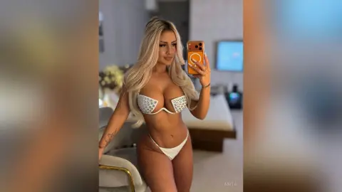 Anita poses for a mirror selfie wearing a white sparkly bikini or lingerie set with long blonde hair.