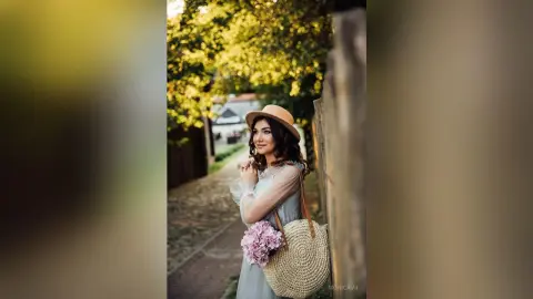 Smiling woman in a blue dress and sun hat carrying a straw bag in a sunny garden setting.