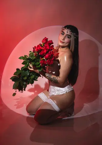 Glamorous woman in white lace lingerie holding a large bouquet of red roses against a pink studio backdrop.