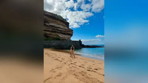 EvaMuskin stands on a sandy beach wearing a black bodysuit, with ocean waves and large rock cliffs behind her.