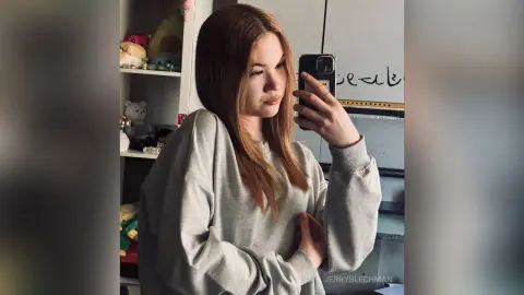 Young woman in a grey sweatshirt taking a casual mirror selfie in a cozy bedroom setting.