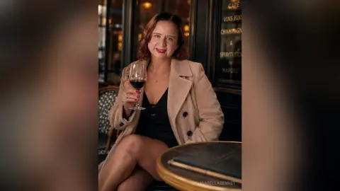 IsabellaBennet wearing a tan coat and black top, holding a glass of red wine while sitting at a cafe table.