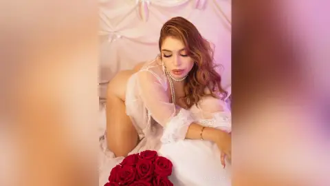 NaiaCollins poses naked against white fabric, wearing a white lace bra and pearl necklace while leaning over white pillows near red roses.