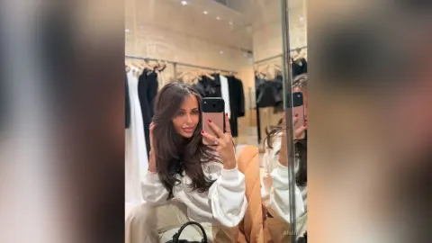 Brunette woman in a white cozy sweater taking a mirror selfie in a bright clothing boutique.