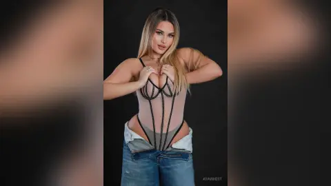 Blonde woman in a sheer black bodysuit and denim jeans posing against a dark studio background.