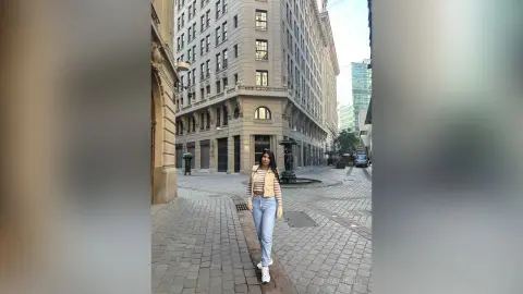 Stylish woman in a beige cardigan and blue jeans walking on a cobblestone city street near historic buildings.