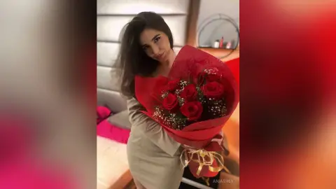 Smiling woman in a beige dress holding a large bouquet of red roses in a cozy indoor setting.