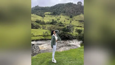 MissyJohansson stands in a green field by a rushing river, wearing a green jacket, white top, and white pants with her hair down.