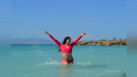 Happy woman in a pink long-sleeve swimsuit splashing in turquoise ocean water under a clear blue sky.