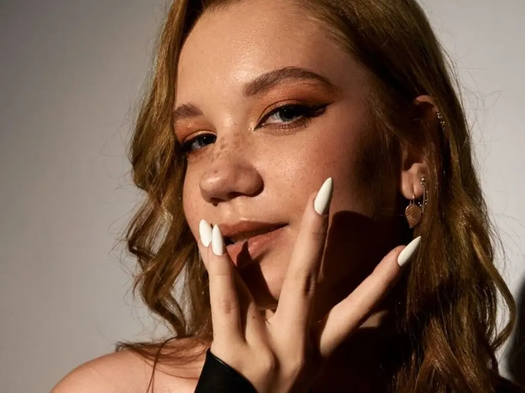 Close-up portrait of a freckled woman with winged eyeliner and long white nails in warm studio lighting.