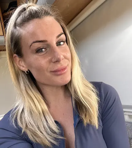 PixiePlayful_ox smiles at the camera with her blonde hair partially pulled back, wearing a blue zip-up top in a bright indoor setting.
