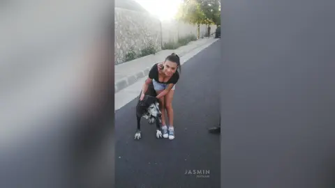 Smiling young woman in a black t-shirt and denim shorts sitting with a black and white dog on a sunny street.