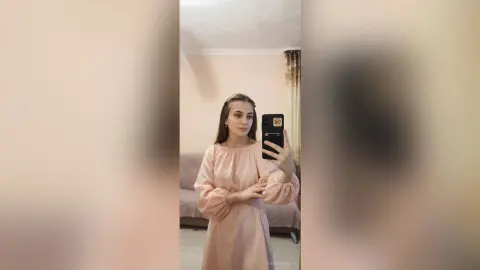 Young woman in a blush pink long-sleeve dress taking a mirror selfie in a cozy, minimalist room.