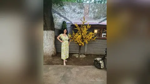 Woman in a yellow floral sundress standing by a blooming yellow tree in a sunny outdoor garden setting.