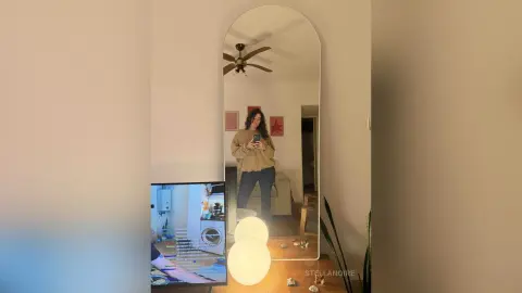 StellaNoire takes a mirror selfie in a tan long-sleeve shirt and dark pants, standing in a living room with a ceiling fan and a TV visible.