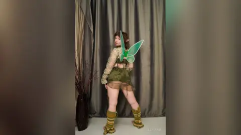 Woman in green fairy costume with wings and boots posing in a studio setting with a whimsical mood.