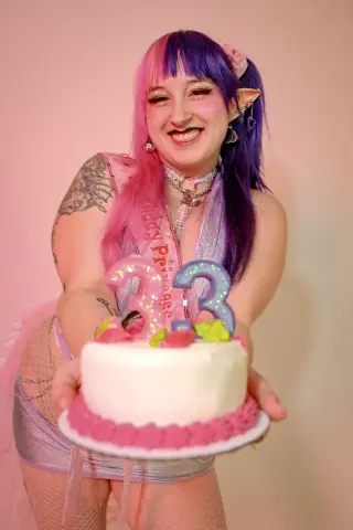 Smiling woman with pink and purple hair in a sparkly outfit holding a birthday cake with number 33 candles.