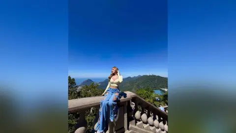Fashionable woman in light yellow top and ripped blue jeans posing against a scenic mountain landscape under a blue sky.
