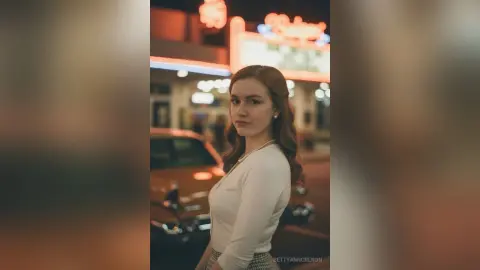 Redheaded woman in a white blouse standing in front of a vintage neon diner at night.