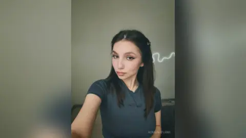 Young woman with long dark hair wearing a casual grey t-shirt in a minimalist indoor setting.