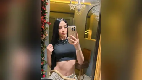 Young woman with long dark hair in a black crop top taking a mirror selfie in a luxury bedroom.