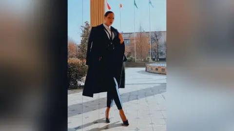 Elegant woman in a long black coat and trousers standing in a sunny urban plaza.