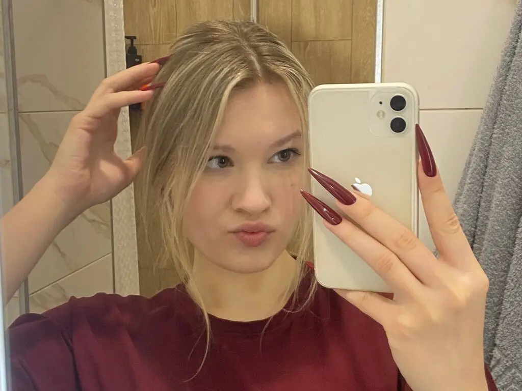 Blonde woman in a maroon shirt taking a mirror selfie with long red nails in a bright bathroom.