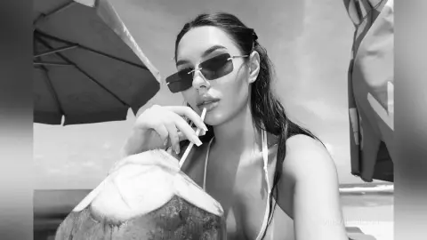 Stylish woman in sunglasses sipping from a coconut under a beach umbrella on a sunny summer day.