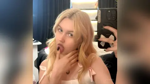 LillyEyes sticks her tongue out while touching her chin, wearing a white top with pink bows and holding a black phone in a mirror selfie.